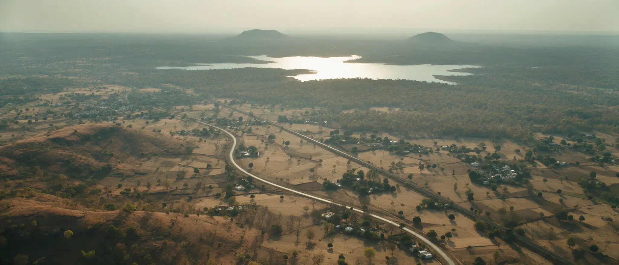 Aerial view over the Vidarbha plains: a road curving between fields towards a lake and the forest beyond